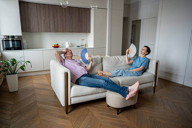 Man and woman fanning themselves on sofa in modern apartment due to air conditioner not cooling properly in summer heat